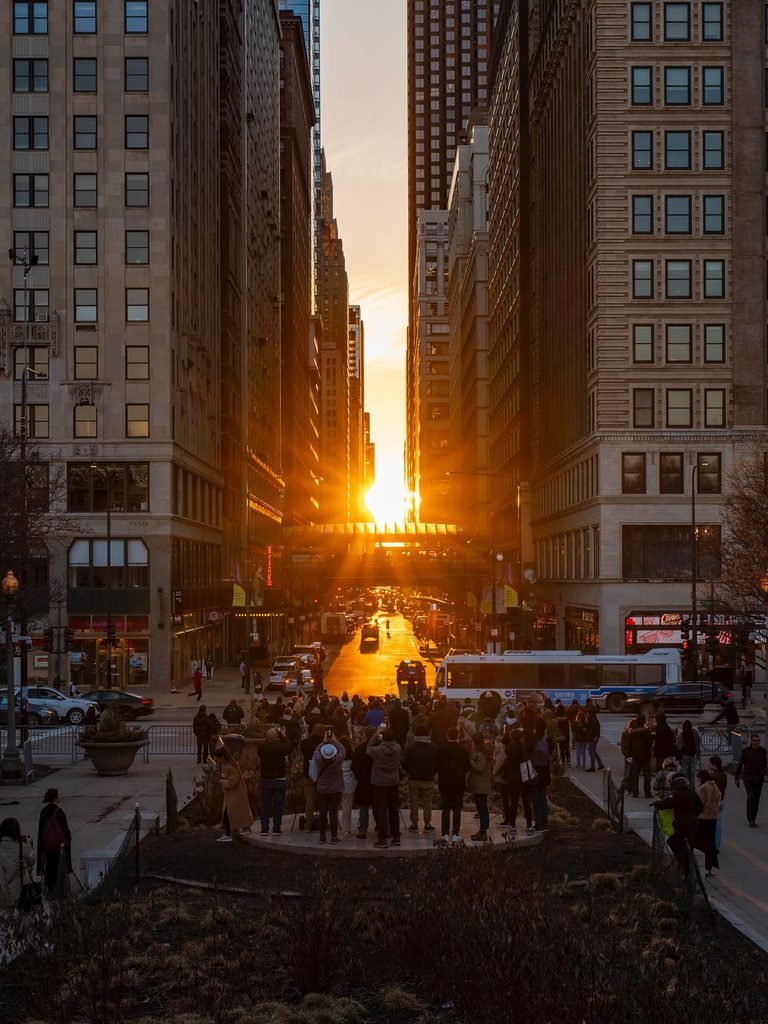 A crowd of spectators gathers to photograph Chicagohenge as the setting sun aligns perfectly between towering downtown skyscrapers.