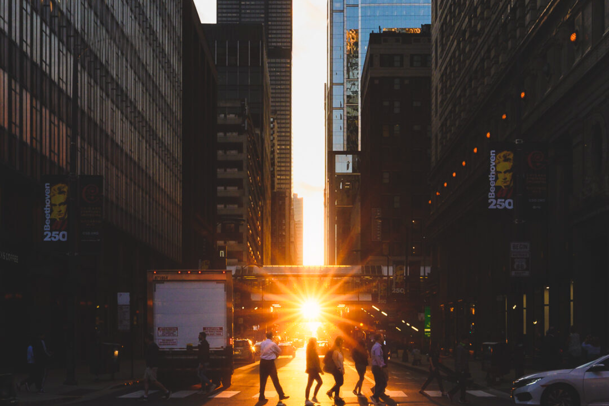 Bright golden sunset aligns between tall Chicago skyscrapers during Chicagohenge while pedestrians cross a shadowy street in silhouette.