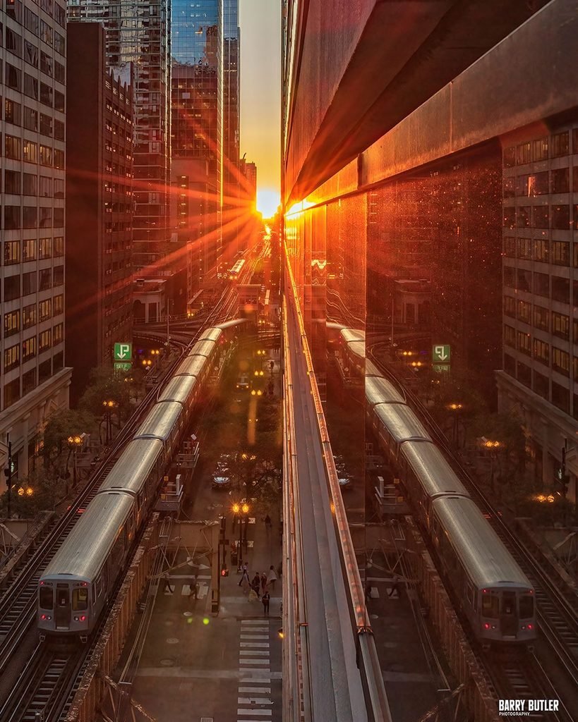 A dramatic golden sunburst flares between Chicago skyscrapers, creating a vivid reflection on glass windows beside elevated CTA L train tracks.