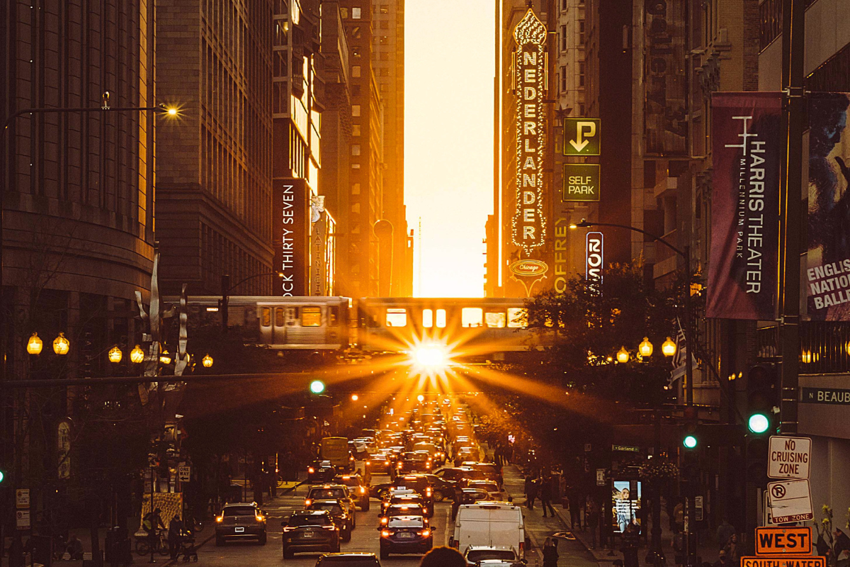 Golden sunset shining down a Chicago street canyon between skyscrapers, illuminating traffic and an elevated train during Chicagohenge.