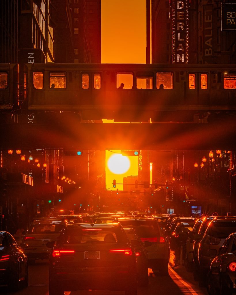Vibrant golden sunburst aligns perfectly between a canyon of Chicago skyscrapers, blazing through a passing elevated L train with passengers and above a busy street with cars and red taillights. Nederlander sign is visible.
