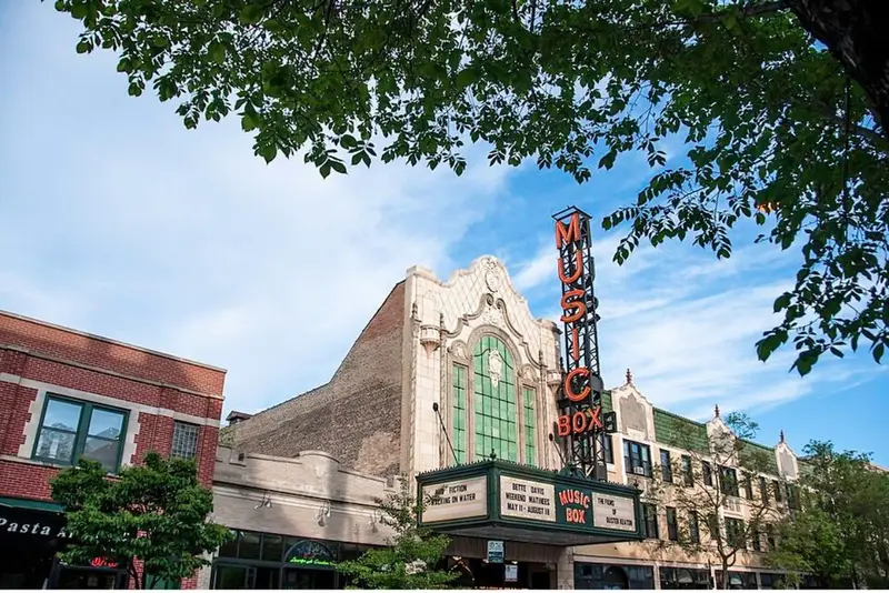 The historic Music Box Theatre in Chicago on a sunny day, featuring its iconic neon sign, marquee, and ornate facade framed by green tree branches.