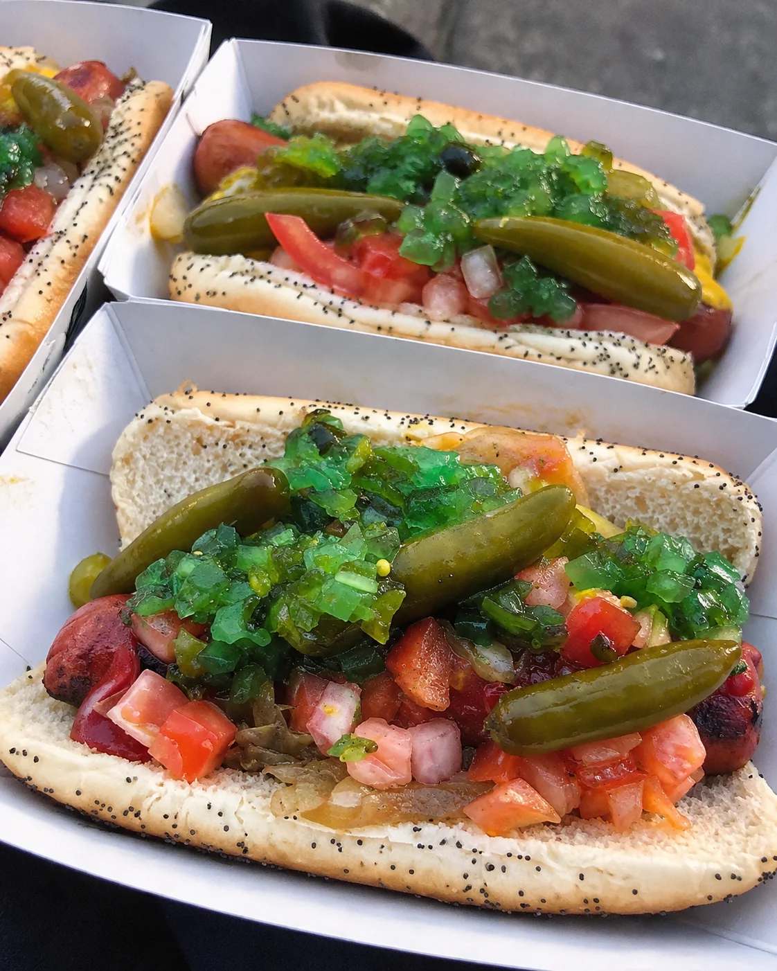 Three fully loaded Chicago-style hot dogs rest in white paper trays, showcasing a variety of colorful toppings inside soft poppy seed buns. Each grilled hot dog is generously piled high with neon green relish, diced red tomatoes, chopped white onions, and whole green sport peppers.