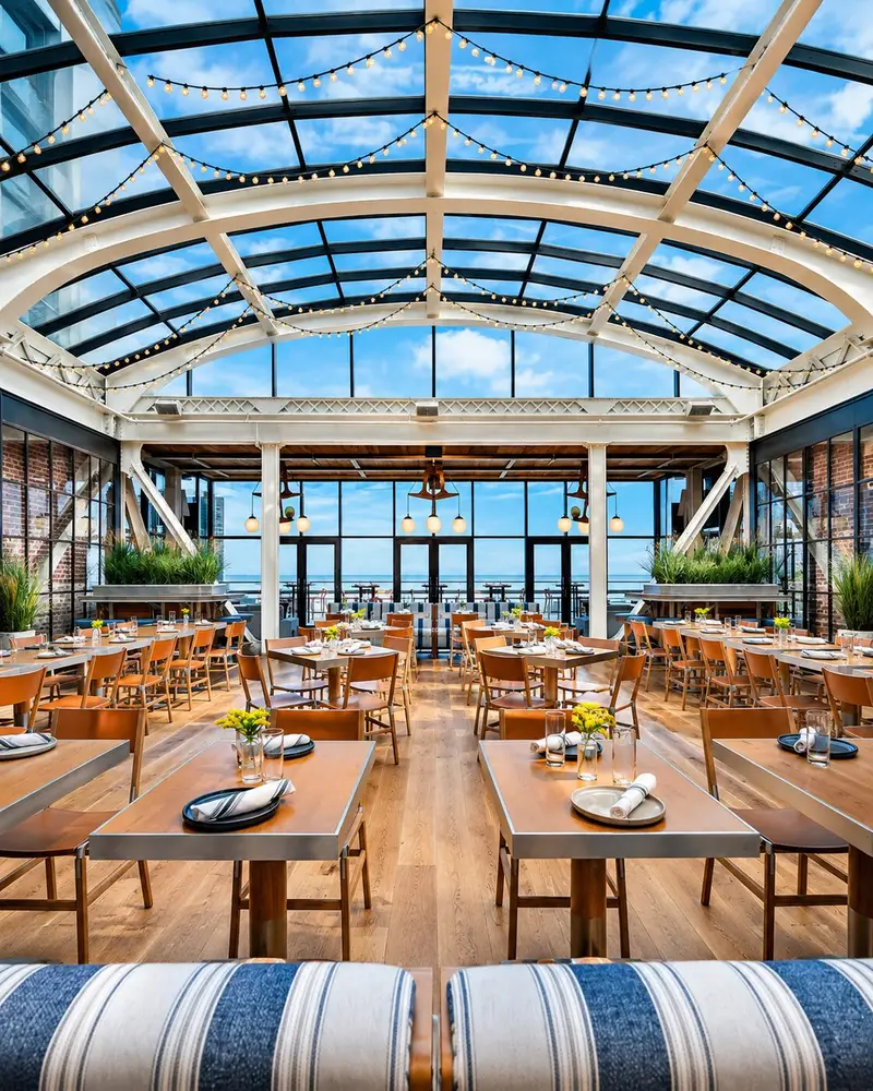 A vibrant rooftop restaurant features a glass-vaulted atrium ceiling, warm wood paneling, and an array of pendant globe lights dangling over the dining area. Blue-and-white striped banquette seating faces wooden tables with tan leather chairs, while a fully stocked bar sits framed by brick pillars in the background.
