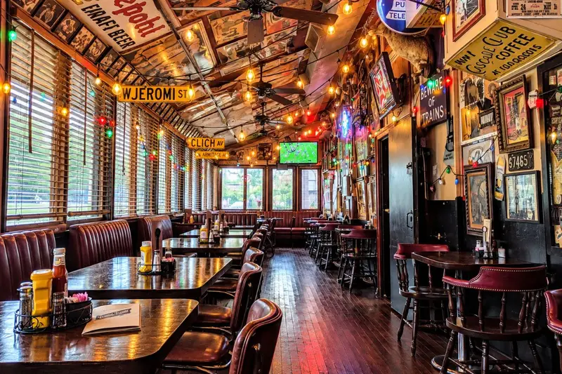 A long, narrow tavern interior features deep red leather booths lined beneath large windows with wooden horizontal blinds on the left. The space is densely decorated with eclectic vintage street signs, neon advertisements, string lights, and historic posters covering the walls and angled ceiling panels.