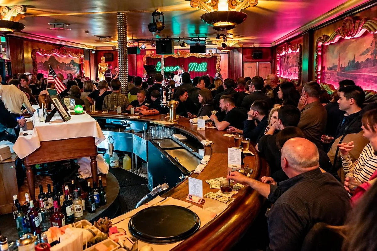 A crowded, dimly lit vintage tavern features patrons packed around a large, curved wooden bar stocked with liquor bottles in the foreground. In the background, a small performance stage glows under warm pink and red ambient lighting, accented by an illuminated "Green Mill" sign.