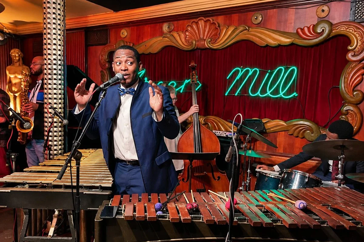 A jazz musician wearing a blue suit and bow tie sings into a microphone while standing behind a vibraphone instrument on a dimly lit stage. In the background, a live band with a saxophonist, double bassist, and drummer performs in front of a red velvet curtain illuminated by a green neon "Green Mill" sign.