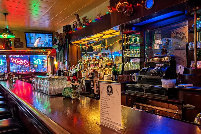 An antique cash register sits on a dark wooden bar counter alongside a printed drink menu and a vase of red roses. The background showcases a fully stocked back bar lit with colorful string lights and neon signs reflecting in a mirror, while rows of clean glassware line the shiny bar top.