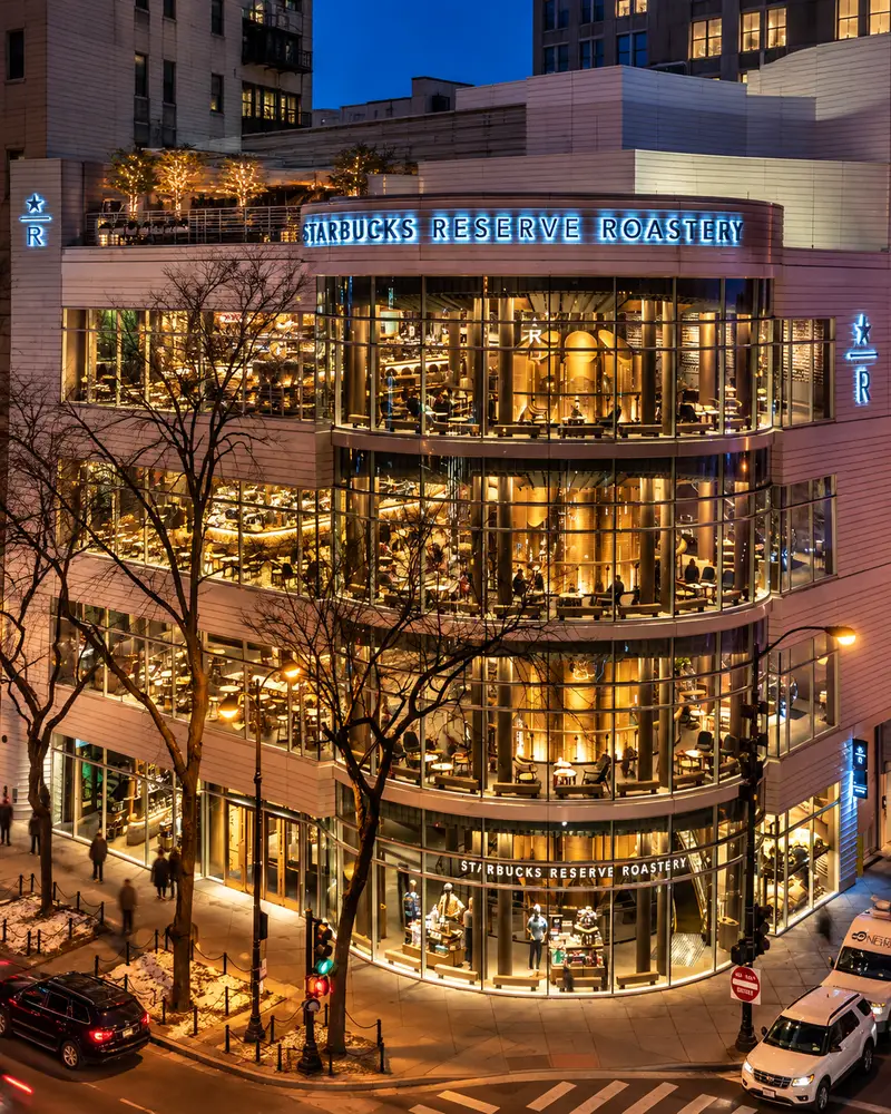 A high-angle evening shot displays the glowing, multi-story glass facade of a modern retail building illuminating a city street corner at twilight. The curved glass exterior reveals a bustling multi-level interior filled with patrons, warm lighting, and a large central roasting cask, while a blue neon sign reads "Starbucks Reserve Roastery" along the top curve.