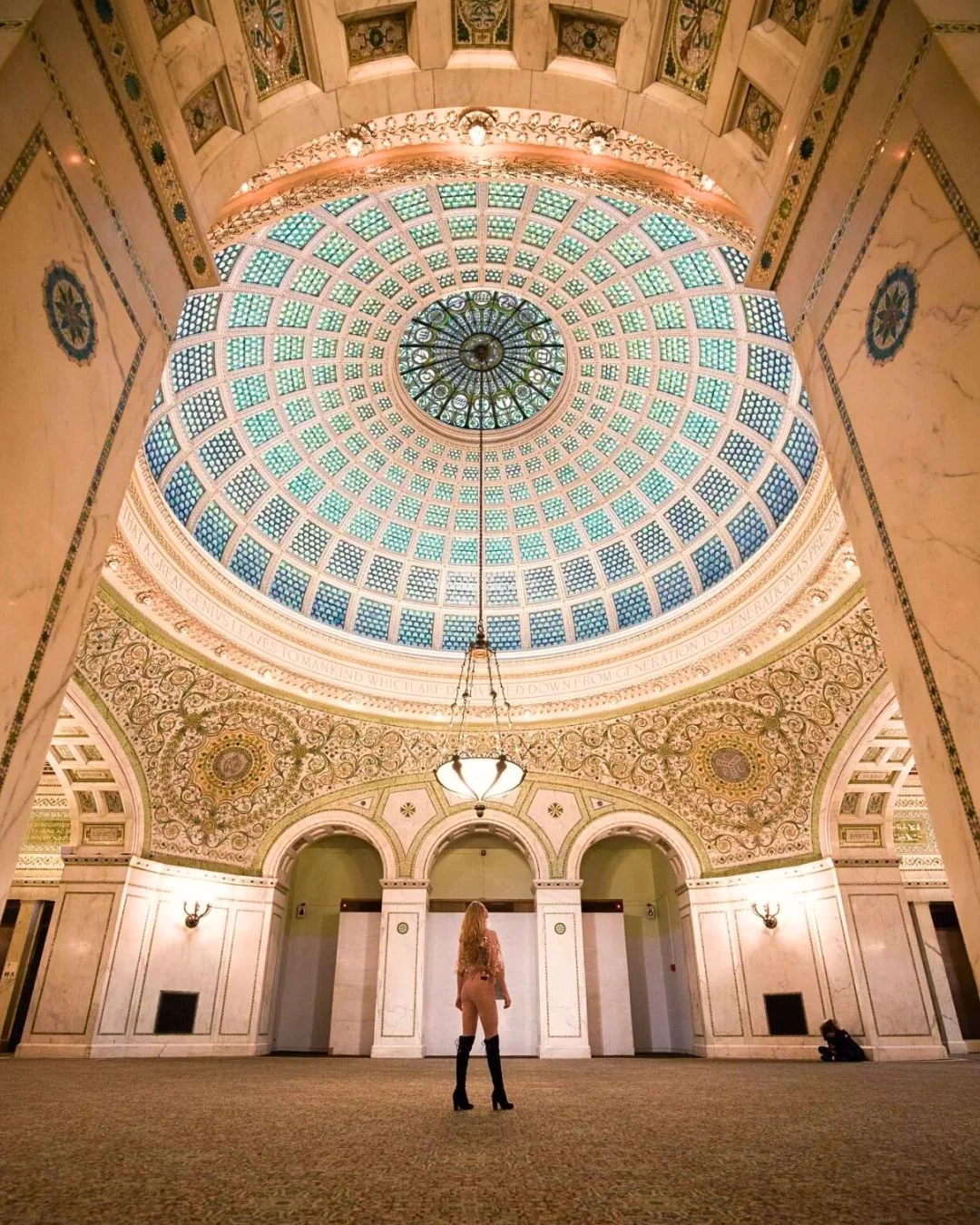 A wide low-angle view highlights a massive, intricately patterned translucent Tiffany glass dome glowing with blue and green tones inside a grand hall. Below the dome, ornate plaster arches, golden mosaics, and a central hanging chandelier frame a single person standing in the middle of the room.