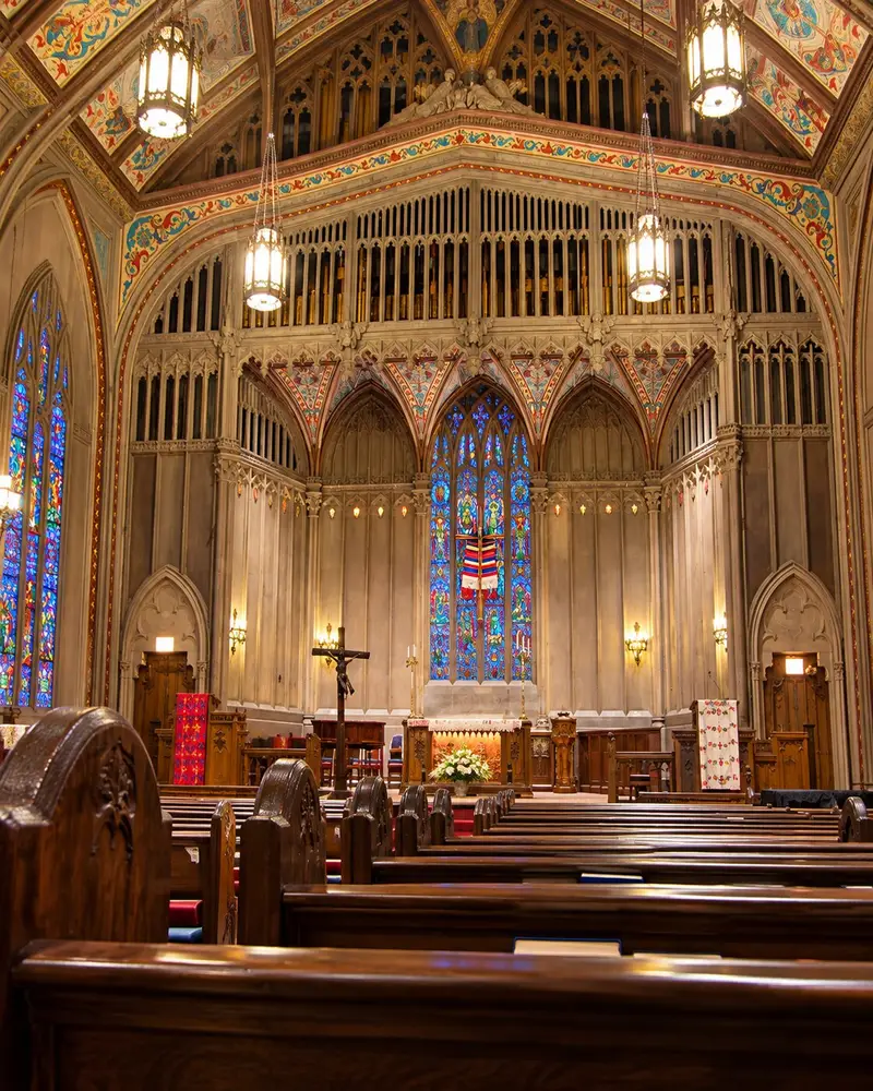 Wooden pews line the center aisle of a majestic gothic-style sanctuary leading toward a raised altar stage. An intricate wooden organ pipe facade towers over a vibrant blue stained-glass window panel, illuminated under the warm glow of hanging brass pendant lanterns.