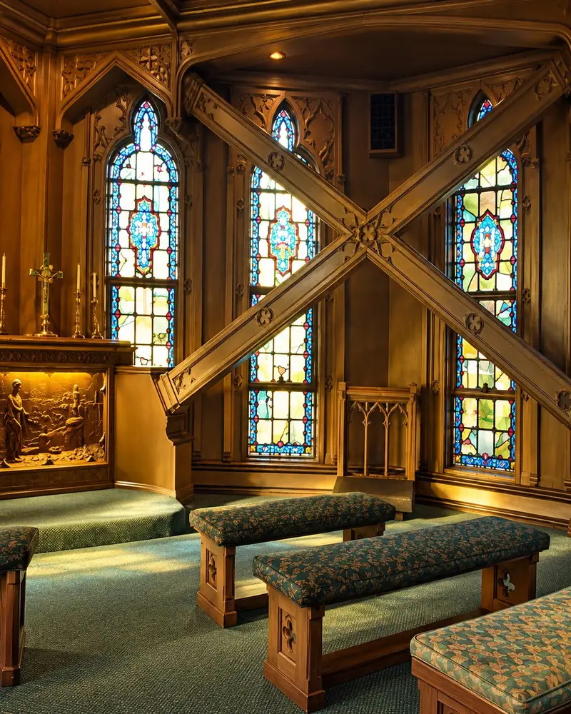 Sunlight streams through vibrant, arched stained-glass windows in an intimate chapel featuring ornate wood-paneled walls and a prominent crisscrossing wooden structural beam. Several cushioned, low-profile benches provide seating on a green carpeted floor, leading towards a small, carved wooden altar on the left.