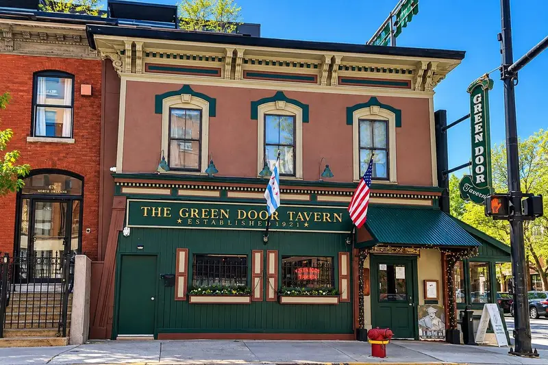 The historic two-story facade of The Green Door Tavern features a deep green painted storefront on the ground level beneath an earth-toned upper story with three double-hung windows. A vertical wooden sign reading "Green Door Tavern" hangs from a black metal bracket near a street corner, accompanied by a hanging American flag and a Chicago city flag over the main sign.