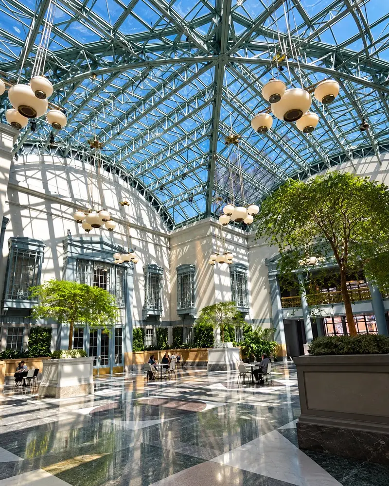Sunlight streams through a massive, vaulted glass ceiling with a teal steel framework into a grand indoor atrium. The bright space features marble tiled floors, large potted trees, hanging sphere-like chandeliers, and visitors sitting at small tables spread throughout the courtyard.
