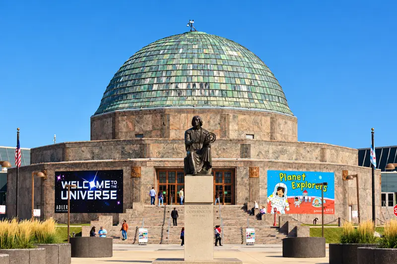 Front entrance of the Adler Planetarium in Chicago on a clear sunny day, featuring the iconic copper dome, the Nicolaus Copernicus monument, and visitors walking up the front steps.