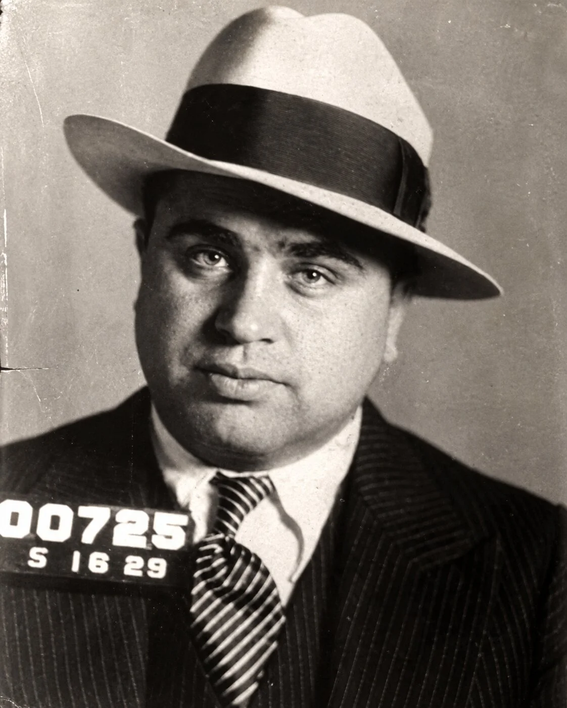 A classic black-and-white mugshot captures American gangster Al Capone looking directly into the camera while wearing a light-colored fedora with a dark band. He is dressed in a pinstripe suit jacket and a striped tie, with identification numbers visible on a placard in the lower left corner.