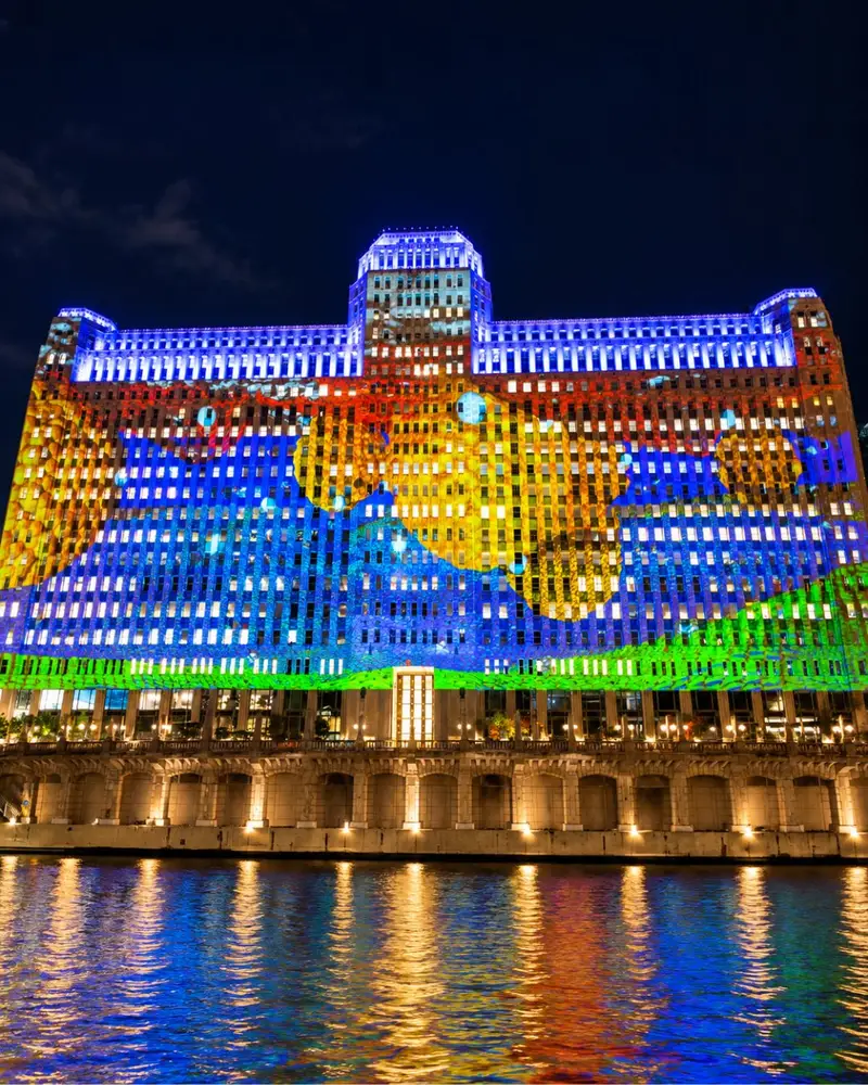 People standing on the Chicago Riverwalk at night watching a massive, colorful digital art projection illuminated across the entire facade of the Merchandise Mart building.