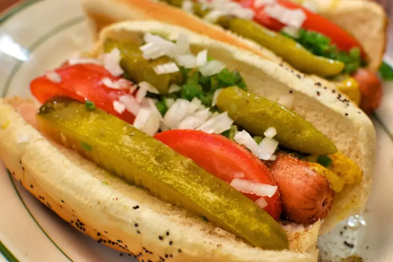A close-up of an authentic Chicago-style hot dog on a poppy seed bun, fully loaded with yellow mustard, green relish, chopped onions, red tomato wedges, a kosher dill pickle spear, sport peppers, and a dash of celery salt.
