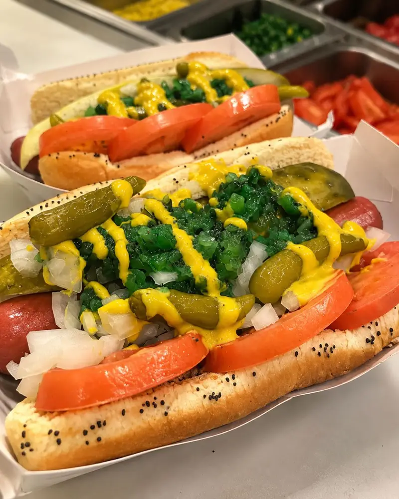 A close-up of an authentic Chicago-style hot dog in a paper tray, loaded with yellow mustard, neon green sweet relish, chopped onions, tomato slices, a pickle spear, and sport peppers on a poppy seed bun.