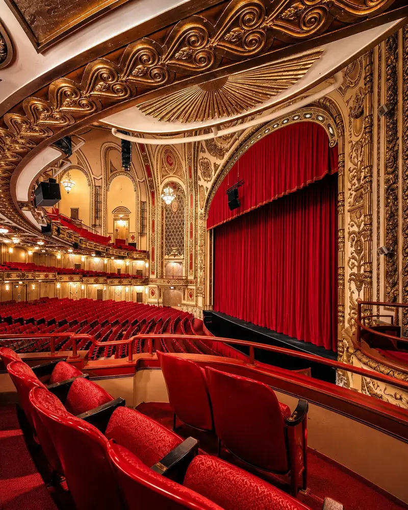 An interior view from an elevated balcony perspective showcases a grand auditorium with curved rows of plush red seats facing a closed red stage curtain. The theater boasts opulent, gold-gilded Neo-Baroque architectural accents, intricately carved details along the balcony trim, and elaborate ceiling murals.