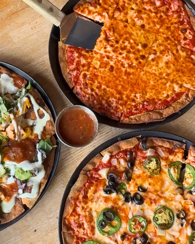 An overhead view of three thin-crust, square-cut tavern-style pizzas on a wooden table, including a classic cheese pie with a metal spatula, a jalapeño and black olive pie, and a specialty taco pizza with a side of salsa.