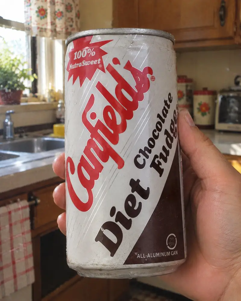 A hand holds up a retro, textured aluminum can of Canfield's Diet Chocolate Fudge soda inside a kitchen setting with a sink and cabinets blurred in the background. The white can features the vintage red cursive Canfield's logo, a red "100% NutraSweet" starburst design, and a dark brown diagonal section highlighting the flavor name.
