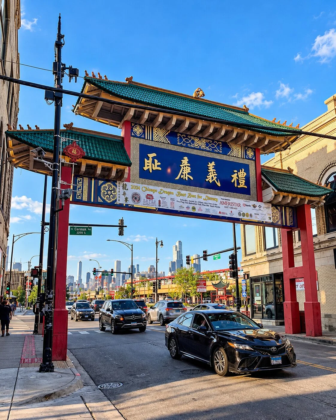A large, traditional Chinese gateway arch with a tiered green roof and intricate blue and red designs spans across a multi-lane city street. Cars travel under the archway, while the distant skyline of Chicago is visible in the background under a clear sky.