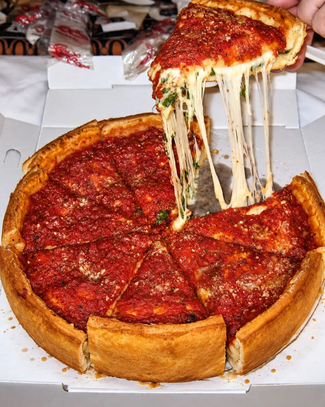 A hand lifts a thick slice of Chicago deep-dish pizza from a cardboard delivery box, creating a dramatic cheese pull of stretched mozzarella. The pizza features a tall, golden-brown crust filled with thick layers of cheese, spinach, and a chunky red tomato sauce seasoned with herbs and parmesan.