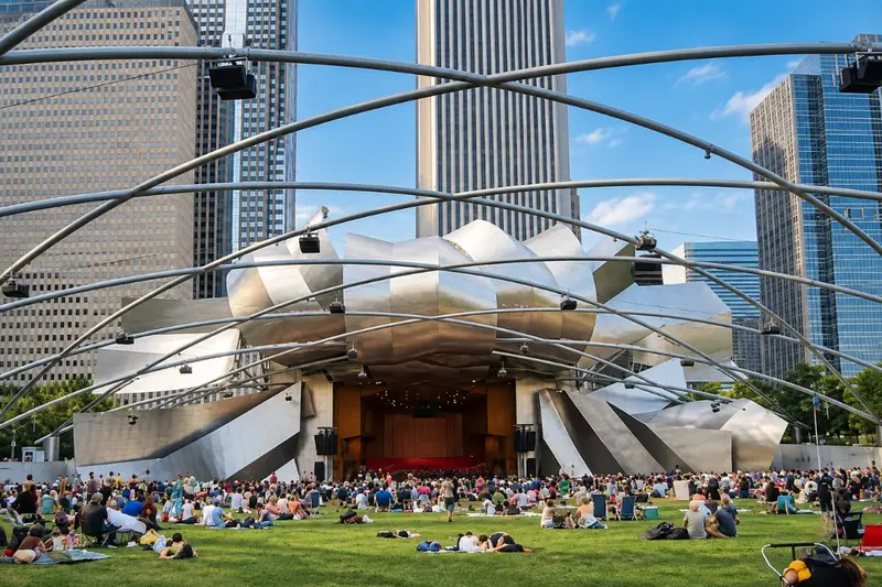 A large, diverse crowd of people sit on the grass enjoying a sunny day in front of the Jay Pritzker Pavilion in Chicago's Millennium Park, with towering skyscrapers in the background.