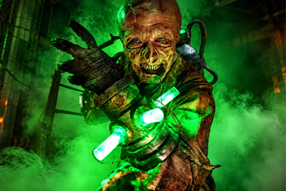 Mutant haunted house scare actor with decaying skin and glowing green chemical gauntlet reaching forward through thick green fog.