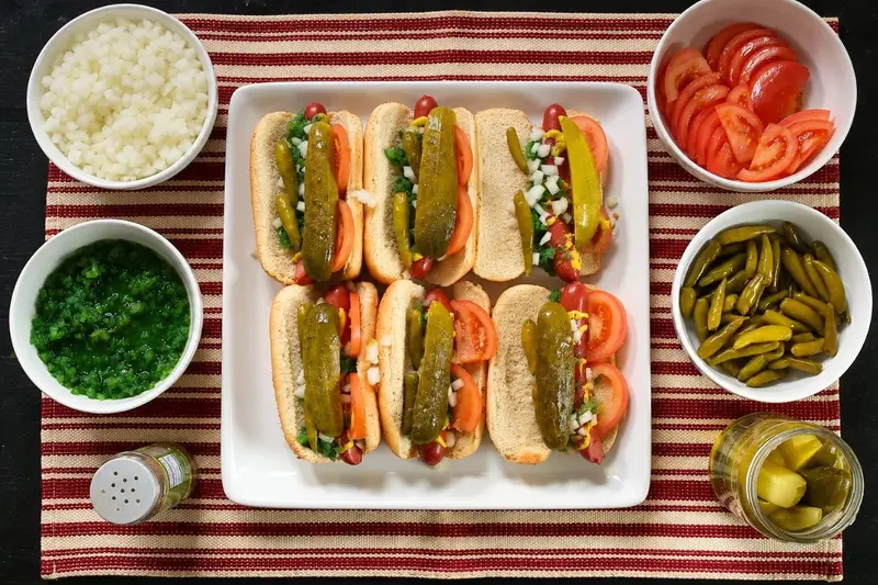 An overhead flat-lay of six hot dogs on a white platter surrounded by individual bowls of chopped onions, green relish, sliced tomatoes, sport peppers, and pickles.