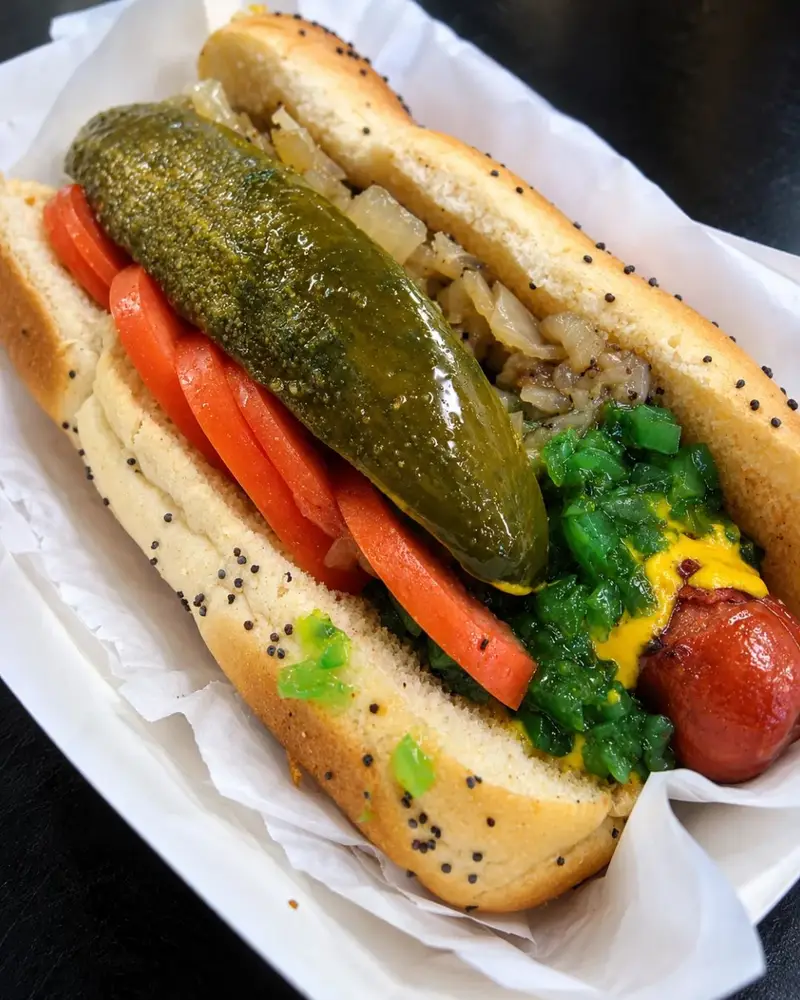A close-up of a Chicago-style hot dog on a poppy seed bun topped with mustard, neon green relish, tomatoes, a large pickle spear, and grilled onions.