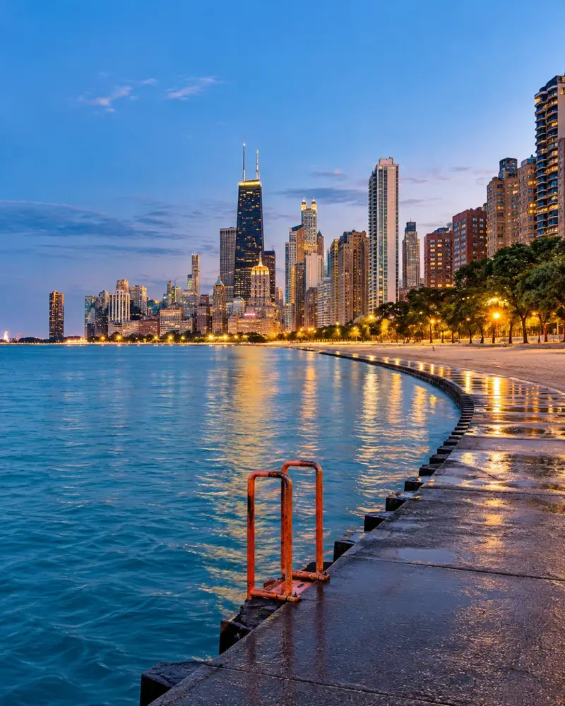A paved walking path curving along the edge of Lake Michigan at dusk, offering a stunning view of the Chicago skyline and John Hancock Center in the distance.