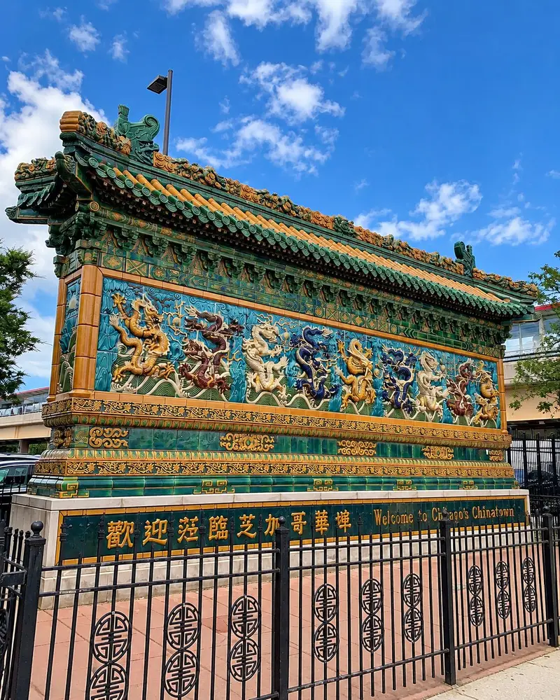 An ornate, freestanding Chinese wall featuring large glazed ceramic tiles with intricate relief carvings of colorful dragons dancing over stylized ocean waves. The structure is capped with a traditional multi-tiered green and orange tiled roof and is enclosed by a black metal fence under a partly cloudy sky.