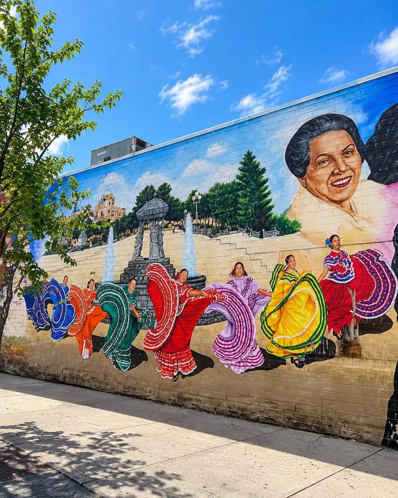 A colorful public street mural painted on a brick building facade showcases a line of women performing traditional Mexican folkloric dances in vibrant, swirling dresses. The dancers are set against a background depicting a historic plaza fountain, with large painted portrait faces flanking the scene under a clear sky.