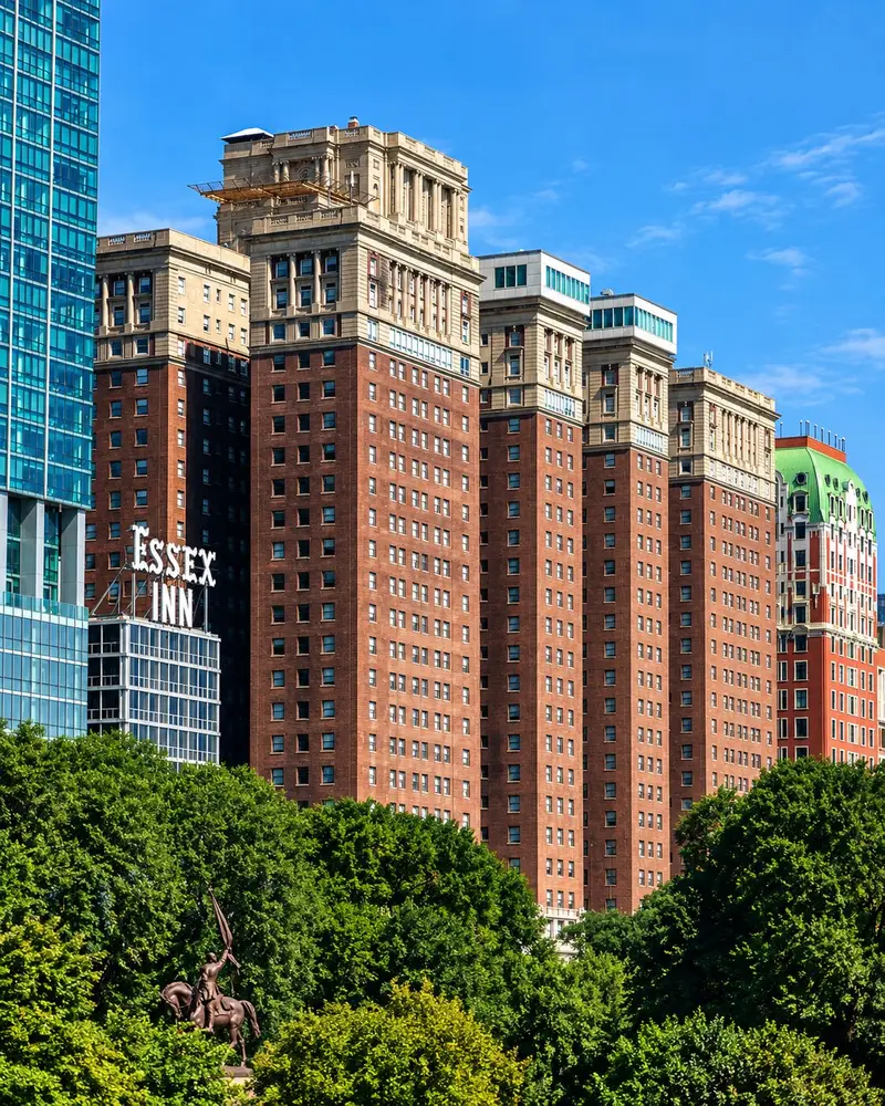 A row of tall, classic brick buildings with ornate stone crowns rises prominently behind a dense line of green trees under a clear blue sky. To the left, a modern teal-glass skyscraper sits beside a structure bearing a white "ESSEX INN" sign, creating a sharp architectural contrast.