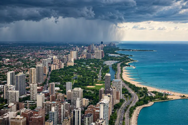 Aerial view of the Chicago skyline, green parks, and Lake Michigan coastline as a massive, dark storm cloud dumps heavy rain over the city.