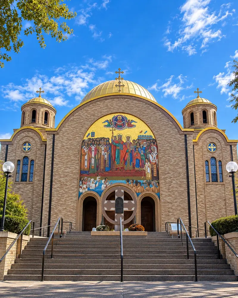 A monumental brick church features a massive golden dome flanked by two smaller domed towers on either side of its front facade. The main entrance is highlighted by concrete stairs leading past a large stone cross toward a giant, intricate gold mosaic mural depicting religious and historical figures.