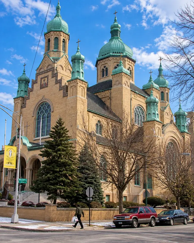 A large historic cathedral built of light-colored brick features numerous prominent, weathered green copper onion domes topped with crosses. The building sits at a street corner behind bare winter trees, where a pedestrian walks on the sidewalk next to parked cars under an overcast sky.