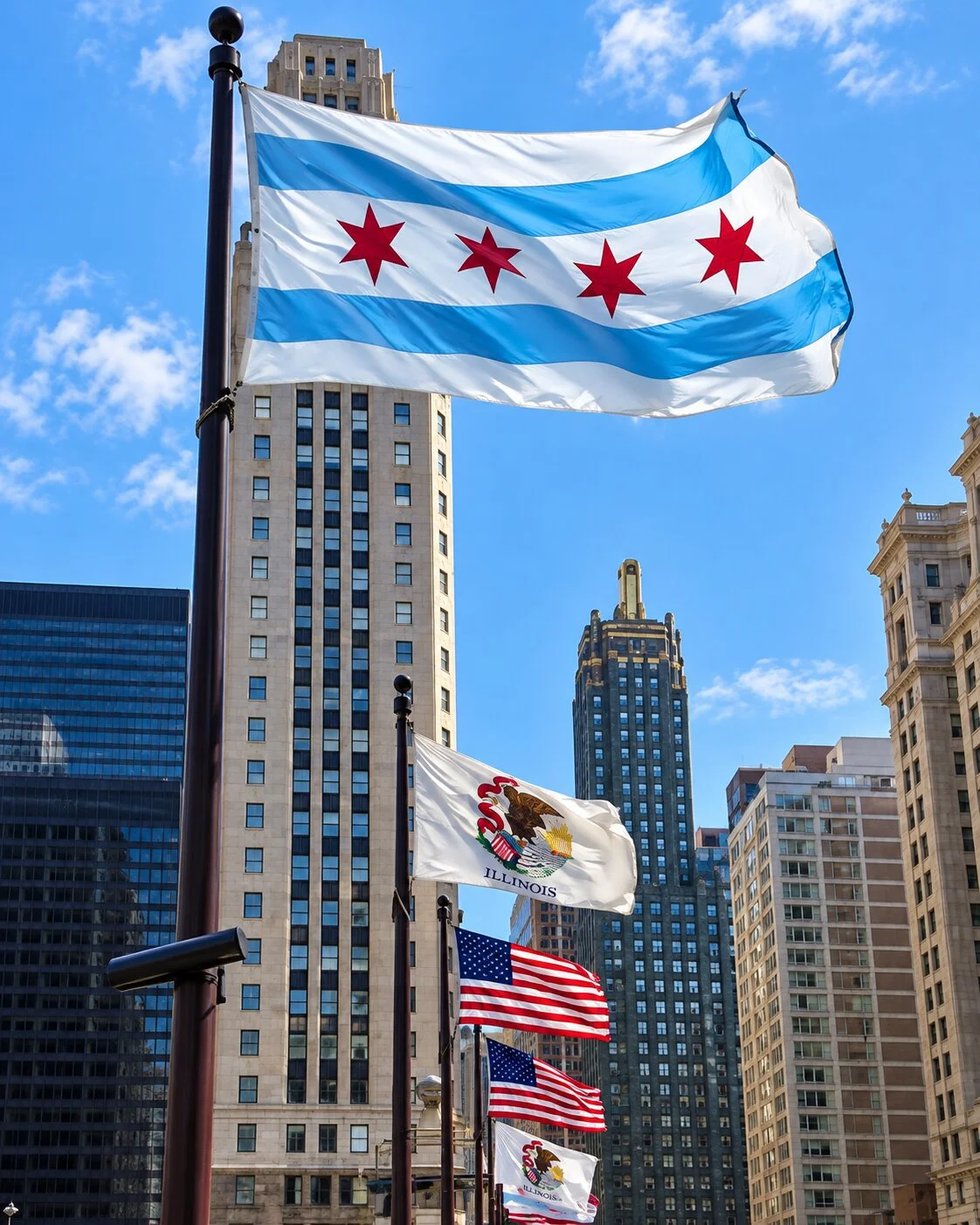 A large city flag of Chicago with two blue stripes and four red six-pointed stars waves prominently on a high flagpole in the foreground. Below it, several other flags including the Illinois state flag and the American flag flutter against a backdrop of dense urban skyscrapers.