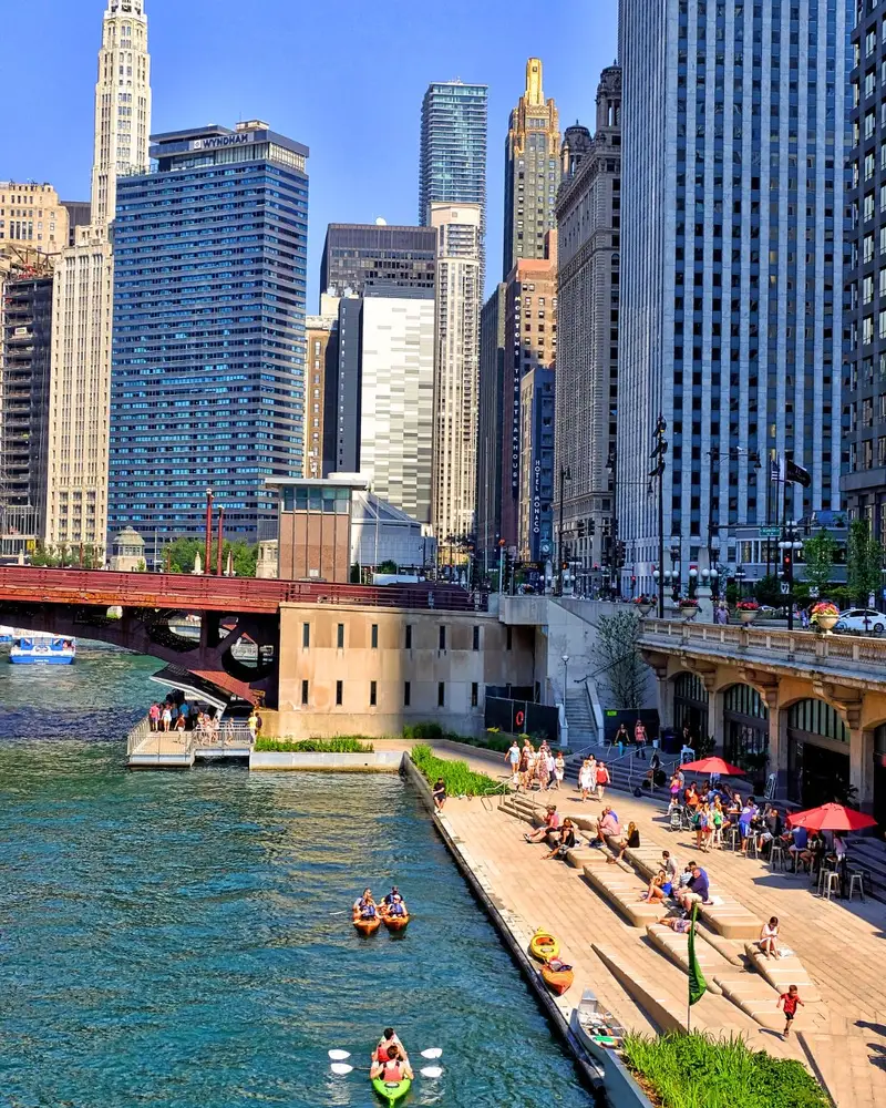 A high-angle shot captures people kayaking on the vibrant teal water of the Chicago River next to a bustling concrete pedestrian walkway. The sunlit scene features multi-tiered concrete seating, patio dining under umbrellas, and an impressive backdrop of towering city skyscrapers.