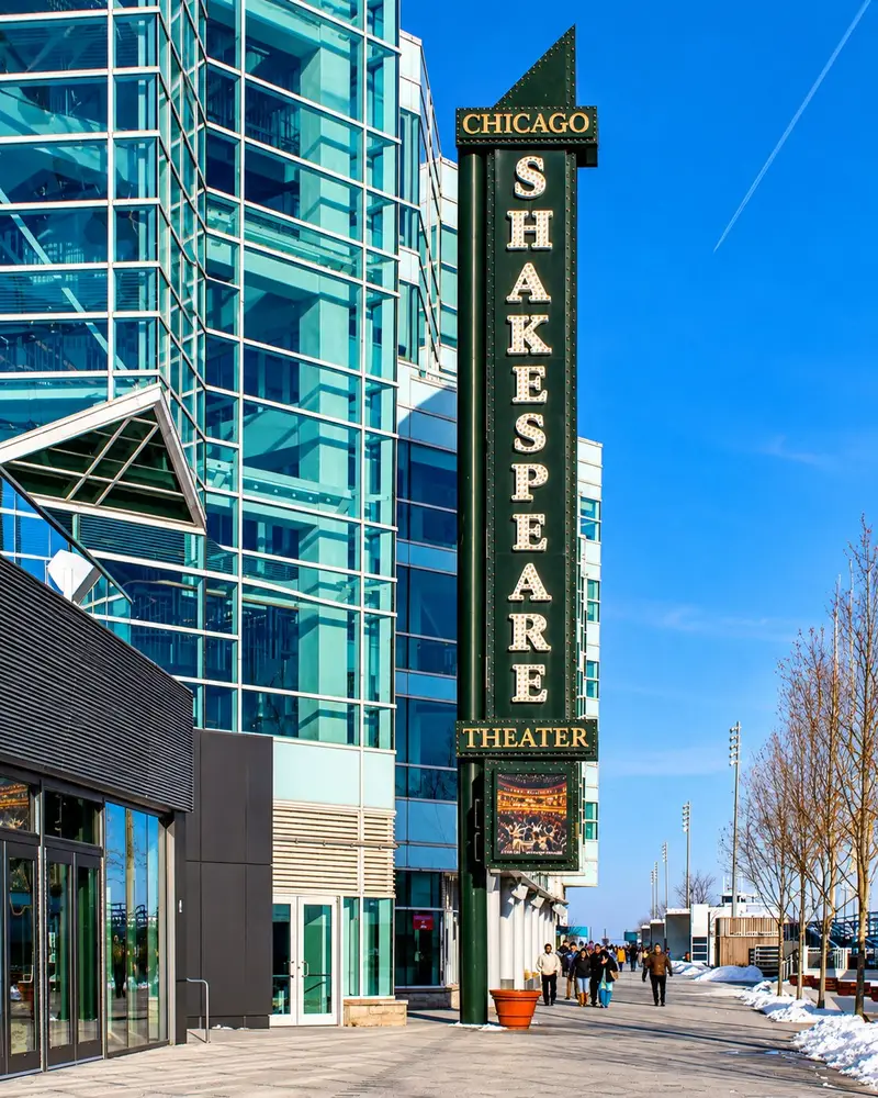 A tall, vertical marquee sign reading "CHICAGO SHAKESPEARE THEATER" stands outside a modern glass-paneled building along a paved walkway. People can be seen walking in the distance under a bright blue sky, with patches of snow lining the sidewalk.