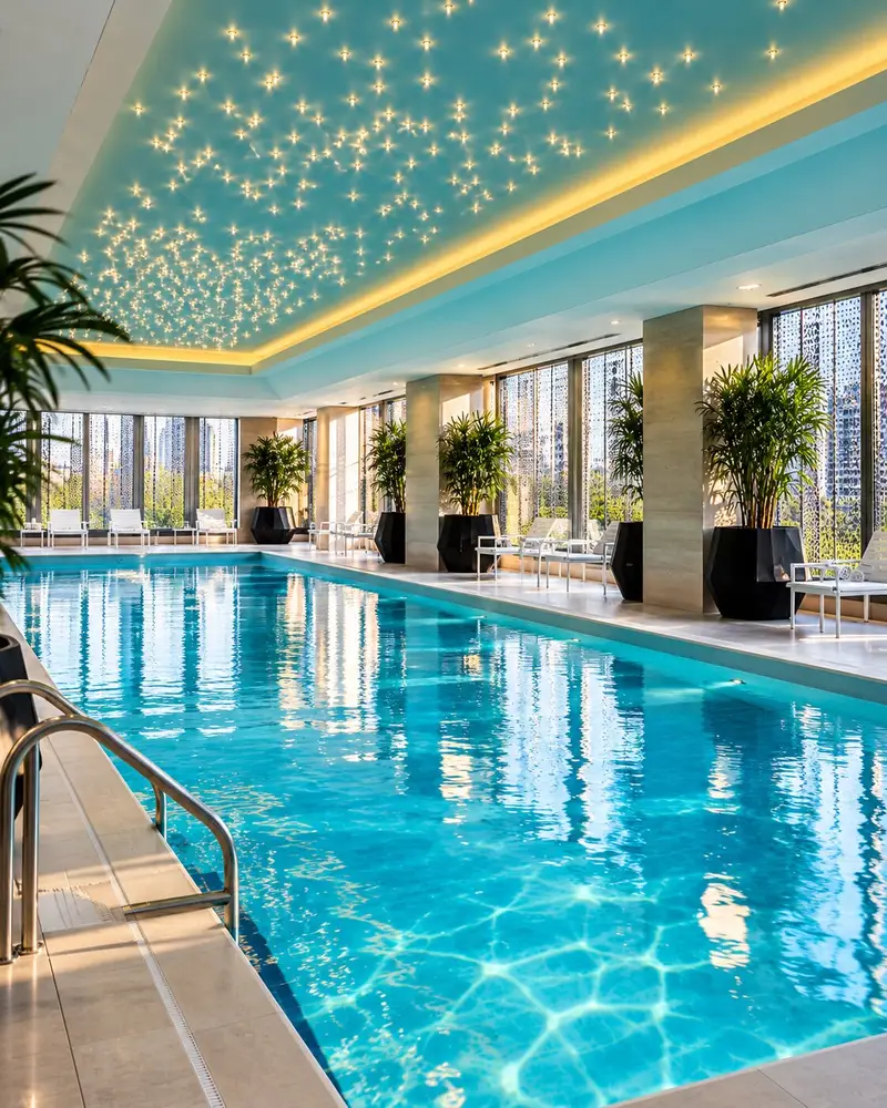 A sleek indoor swimming pool in different angle is filled with glowing turquoise water that reflects a vibrant teal ceiling adorned with an artistic cluster of small, twinkling lights. White lounge chairs and large potted green palms line the clean, minimalist pool deck, which features floor-to-ceiling windows along the far wall.