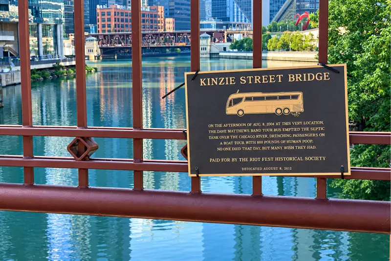 A humorous illustration of a bronze historical plaque on a red bridge railing, commemorating the 2004 Dave Matthews Band incident and credited to the Riot Fest Historical Society.