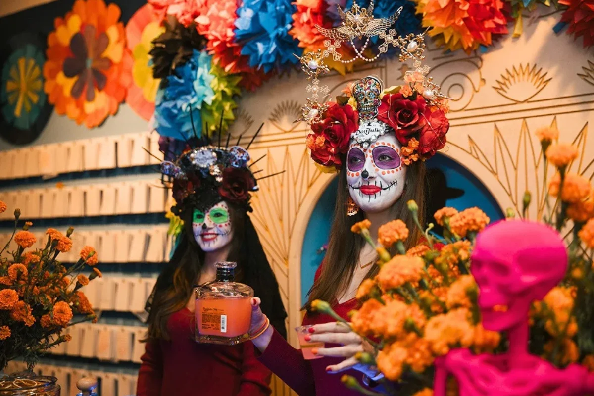 Two women wearing intricate Día de los Muertos sugar skull makeup and elaborate floral crowns stand behind a table decorated with bright orange marigolds and a neon pink skull.