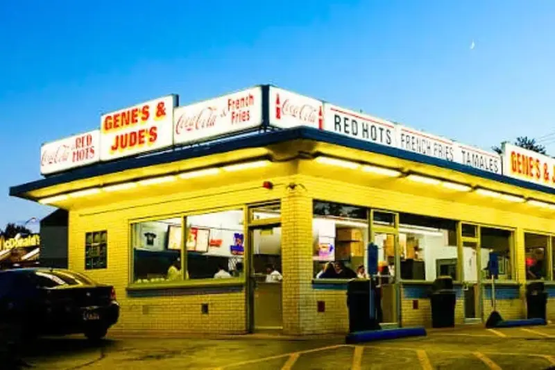 The brightly lit exterior of Gene and Jude's hot dog stand at dusk in River Grove, Illinois, featuring vintage signs for Red Hots and French Fries, with a McDonald's sign visible in the background across the street.