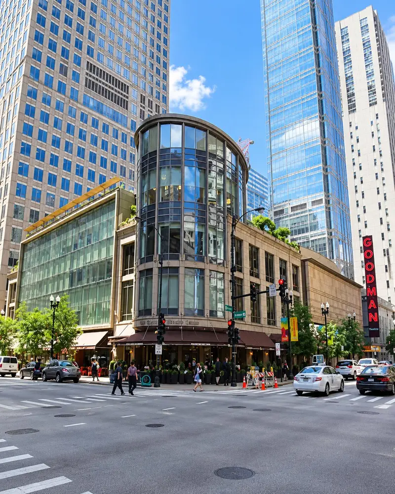 A street-level shot captures a city intersection featuring a multi-story building with a distinctive curved, modern glass rotunda corner. In the background, taller skyscrapers rise into the sky, and a vertical neon sign for the Goodman Theatre is visible on the far right.