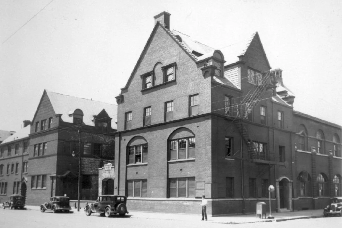 A historic black-and-white photograph of the imposing brick buildings of the Hull-House settlement in Chicago, featuring vintage cars parked along the street.