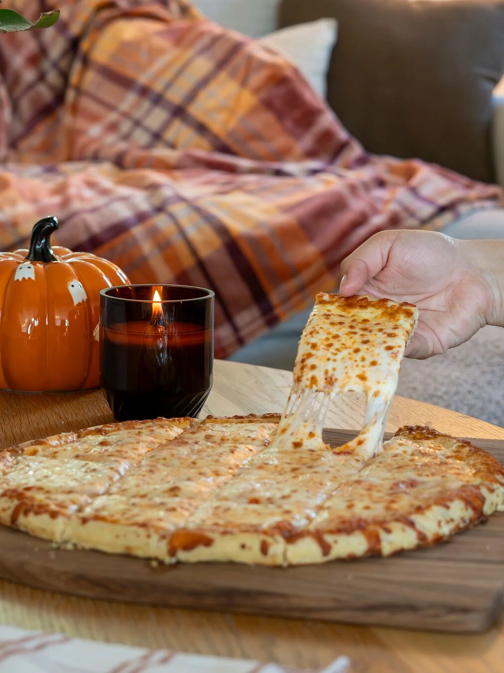 A close-up of a hand pulling a long, melted cheese strand from a piece of tavern-style square-cut cheese pizza on a wooden board. The background features a cozy living room setting with a lit dark glass candle, a ceramic pumpkin painted with ghosts, and a plaid blanket.