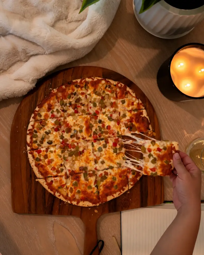 An overhead view of a thin-crust, square-cut Chicago tavern-style pizza on a wooden pizza peel. A hand lifts a center square topped with diced peppers, creating a gooey cheese pull. A glowing three-wick candle and a cozy white blanket sit in the background.